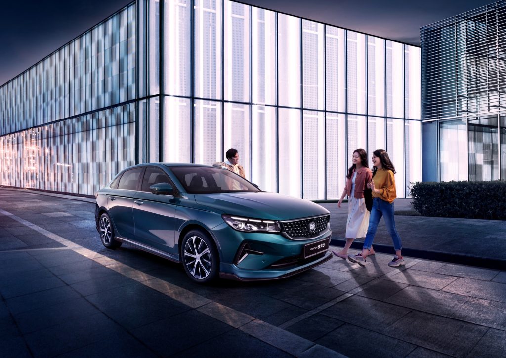 Proton S70 sedan bergaya diparkir di kawasan bandar moden dengan orang muda berjalan di sekelilingnya